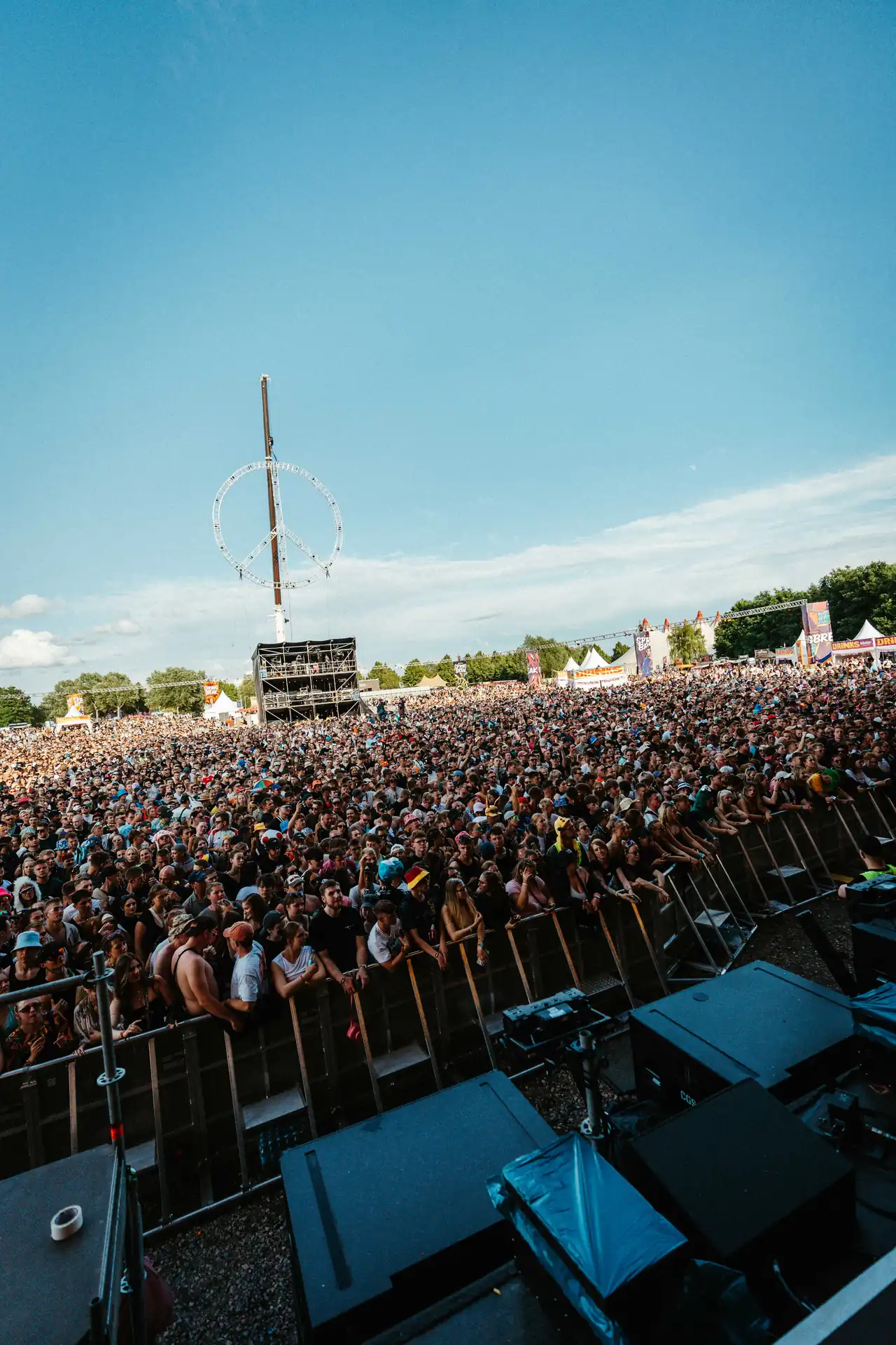 Große Menschenmenge bei einem Outdoor-Musikfestival vor einer Bühne mit klarem blauen Himmel und einem großen Friedenszeichen aus Licht im Hintergrund.