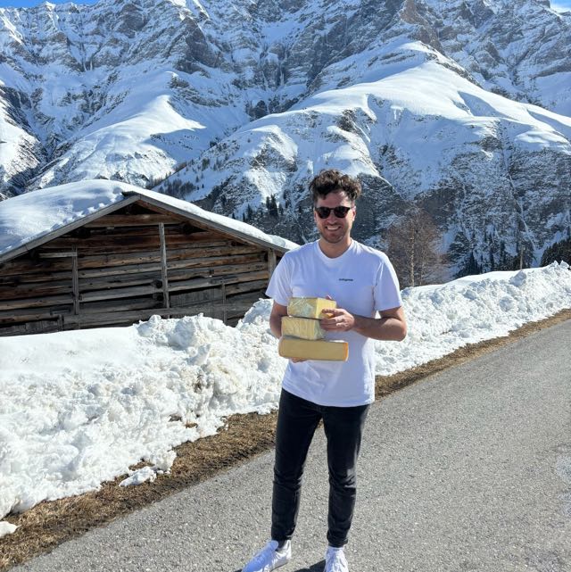Mann mit Sonnenbrille hält drei große Käselaibe stehend auf einer Bergstraße mit schneebedeckten Bergen und einer Holzhütte im Hintergrund.