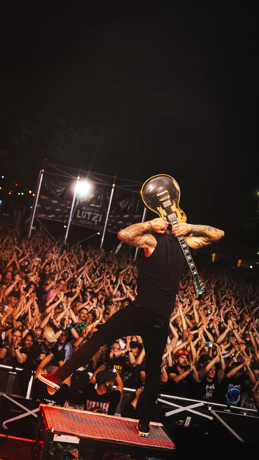 Ein tätowierter Musiker mit schwarzem Outfit und Gitarre hinter dem Kopf steht auf einer Bühne vor einem jubelnden Publikum bei einem Konzert.