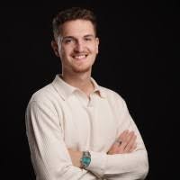 Junger Mann mit kurzen Haaren und hellem Hemd lächelt mit verschränkten Armen vor dunklem Hintergrund.