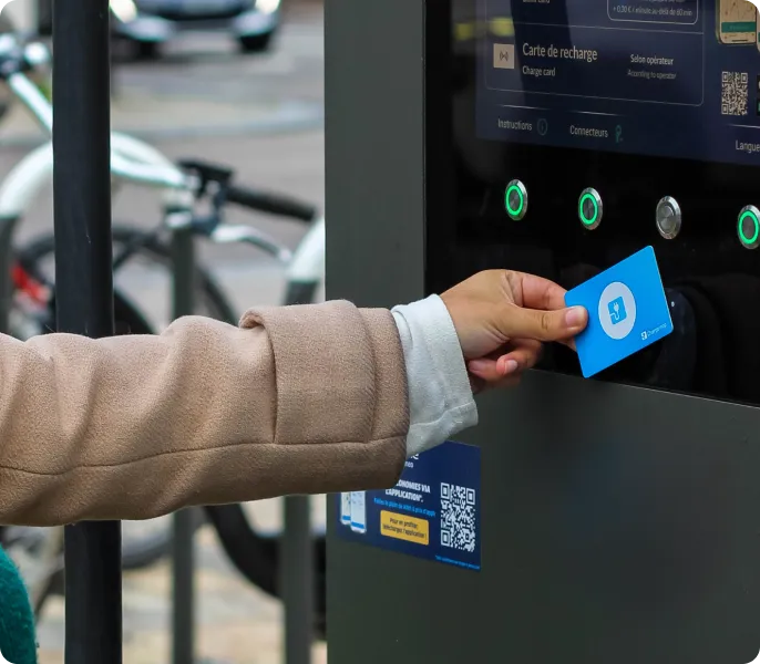 Personne en manteau beige utilisant une carte sans contact bleue sur un distributeur de billets noir à l'extérieur près des bicyclettes.