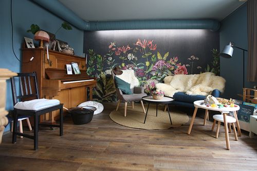 Gemütliches Wohnzimmer mit einem Klavier, einem Stuhl, Sesseln mit Fellüberzügen, einem kleinen runden Teppich, einem Tisch mit Dekoration und einer Wand mit floralem Wandbild.