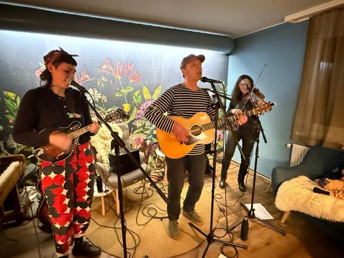Drei Musiker spielen in einem gemütlichen Raum mit Wandbild, eine Frau spielt Mandoline, ein Mann Gitarre und eine Frau Geige.