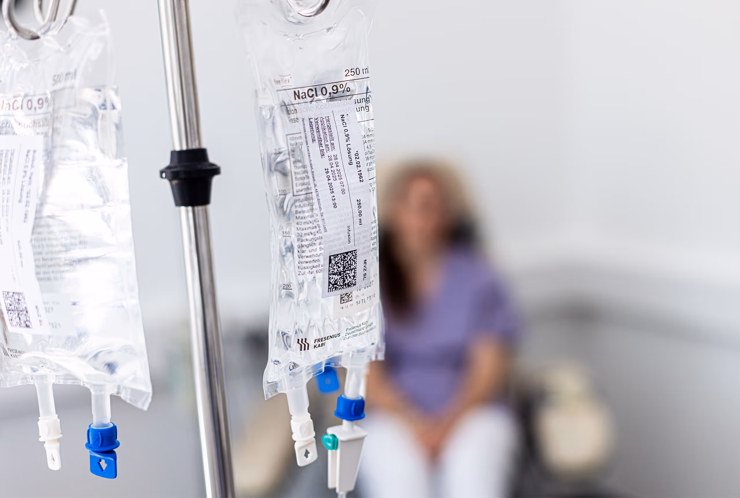 Zwei Infusionsbeutel mit NaCl 0,9% Kochsalzlösung vor einer verschwommenen sitzenden Person.