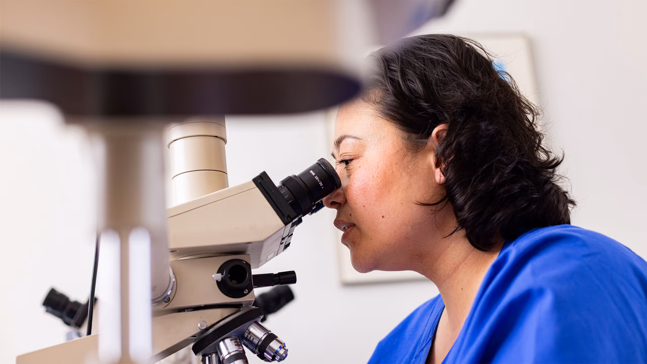 Frau in blauem Labor-Kittel schaut durch ein Mikroskop in einem Labor.