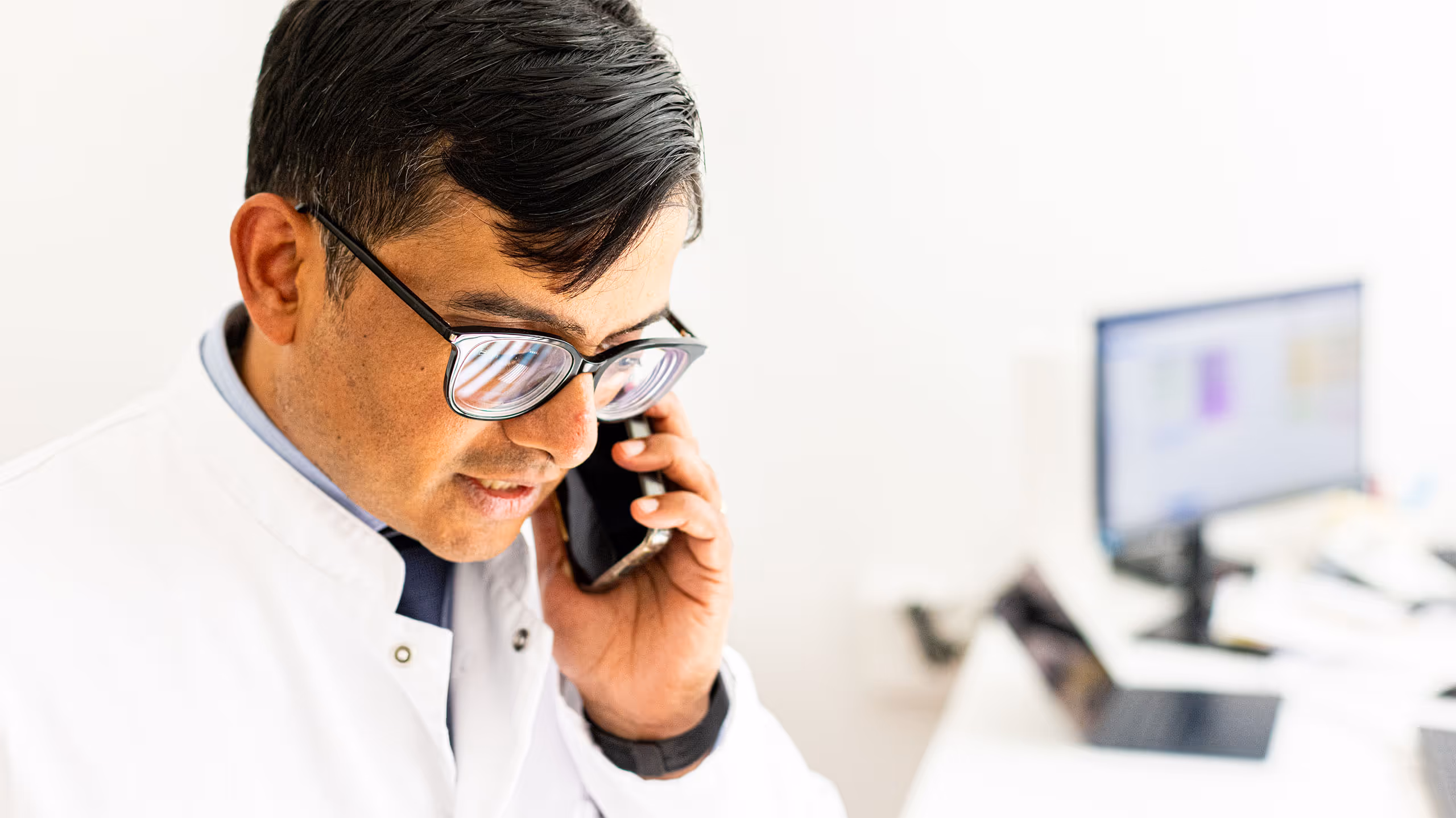 Männlicher Arzt mit Brille im weißen Kittel spricht am Telefon in einem hellen Büro mit Computer im Hintergrund.