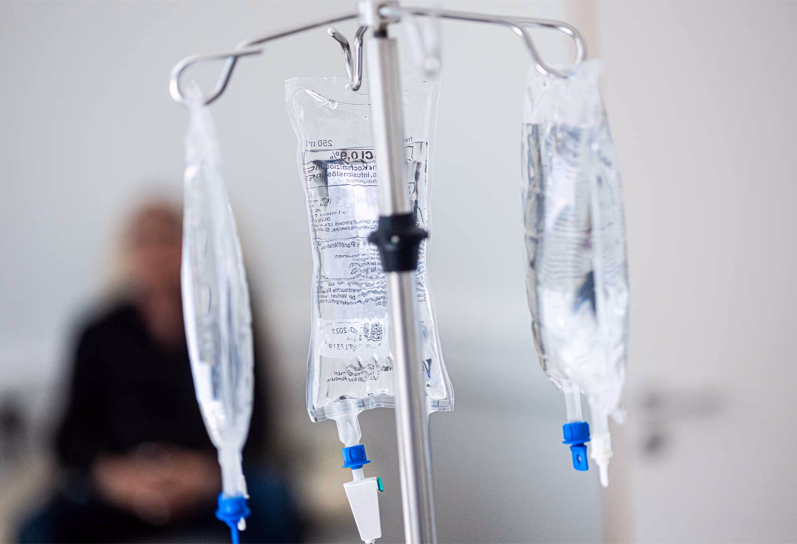 Drei Infusionsbeutel an einem Ständer mit unscharfem Hintergrund eines sitzenden Menschen.