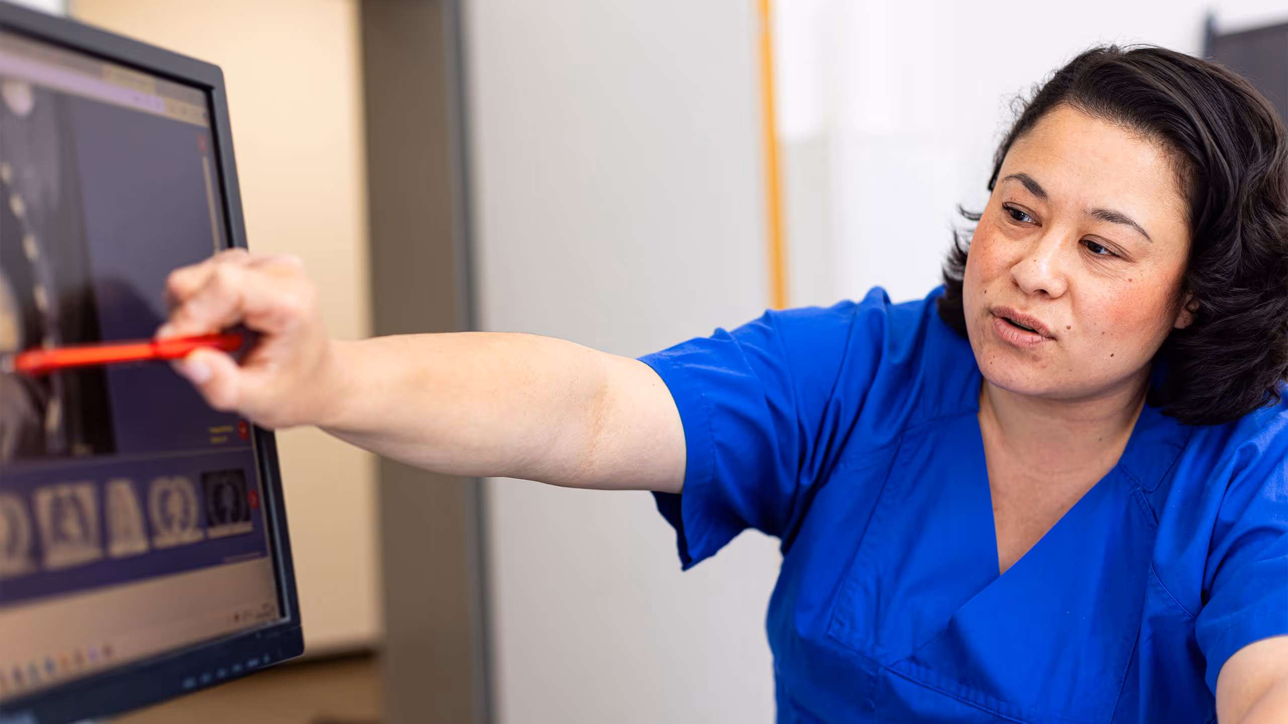 Eine Frau in blauer medizinischer Kleidung zeigt mit einem roten Stift auf einen CT-Scan oder Röntgenbild auf einem Computermonitor.