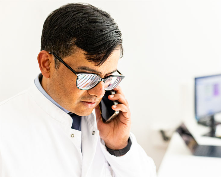 Mann in weißem Kittel mit Brille, der konzentriert telefoniert vor einem Computerbildschirm.