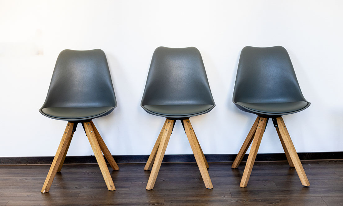Drei dunkelgraue Stühle mit Holzbeinen vor einer weißen Wand auf dunklem Holzboden.