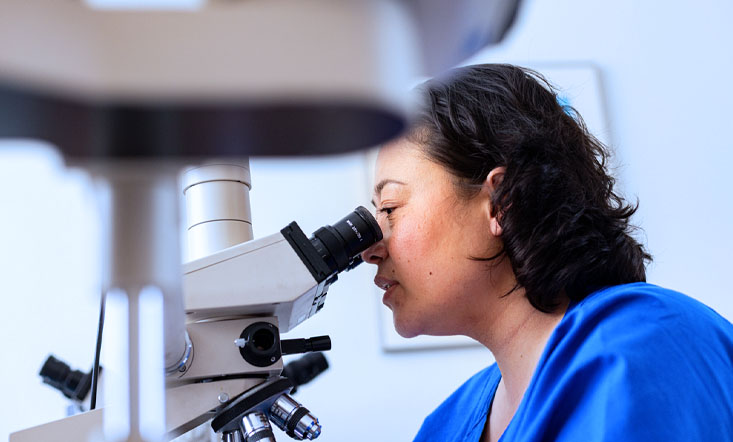 Eine Frau in einem blauen Kittel blickt durch ein Mikroskop in einem Labor.