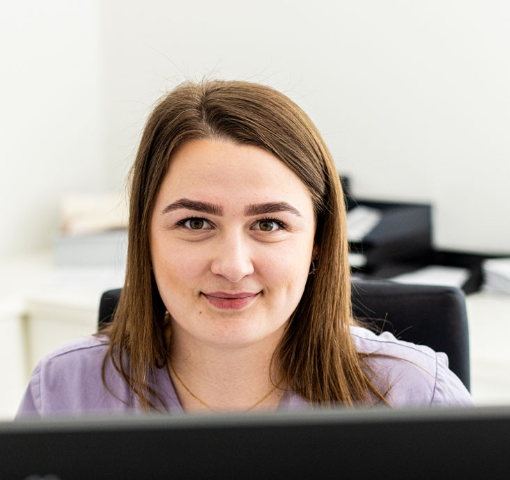 Junge Frau mit braunen Haaren und lila Bluse, die an einem Schreibtisch vor einem Computer sitzt und lächelt.