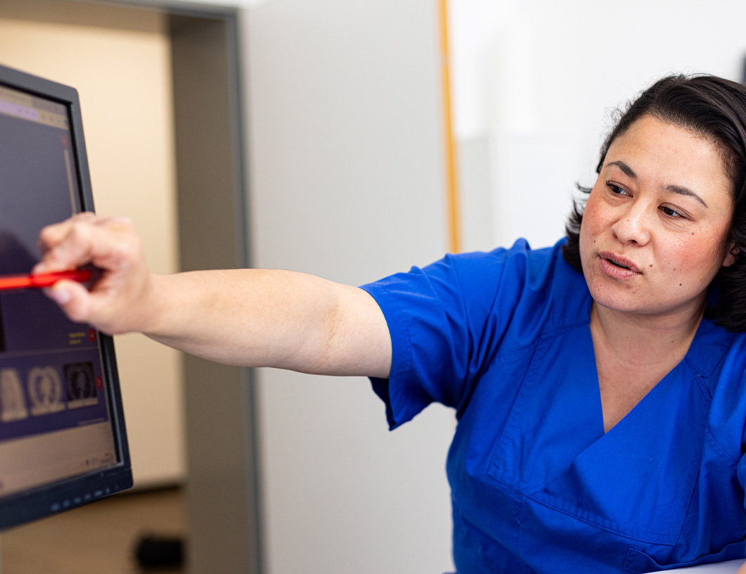Eine medizinische Fachkraft in blauer Arbeitskleidung zeigt mit einem roten Stift auf einen Computermonitor mit medizinischen Bildern.