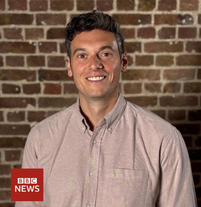 A middle aged white man with dark hair is facing the camera and smiling. In the bottom left is a logo which says BBC News