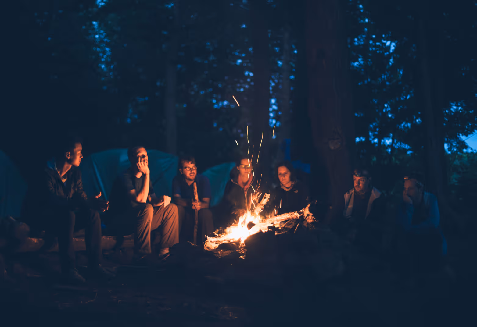 Young adults/youth sitting around a bonfire at night in the woods.