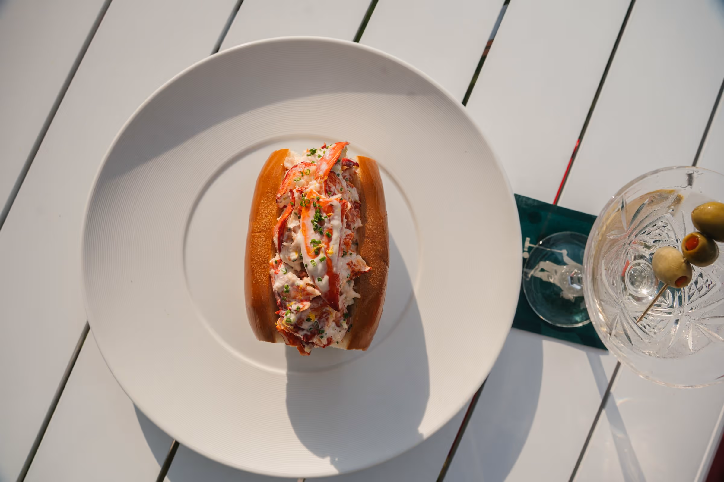 Chilled lobster roll topped with chives on a brioche bun beside a dirty martini with olives, shot from above