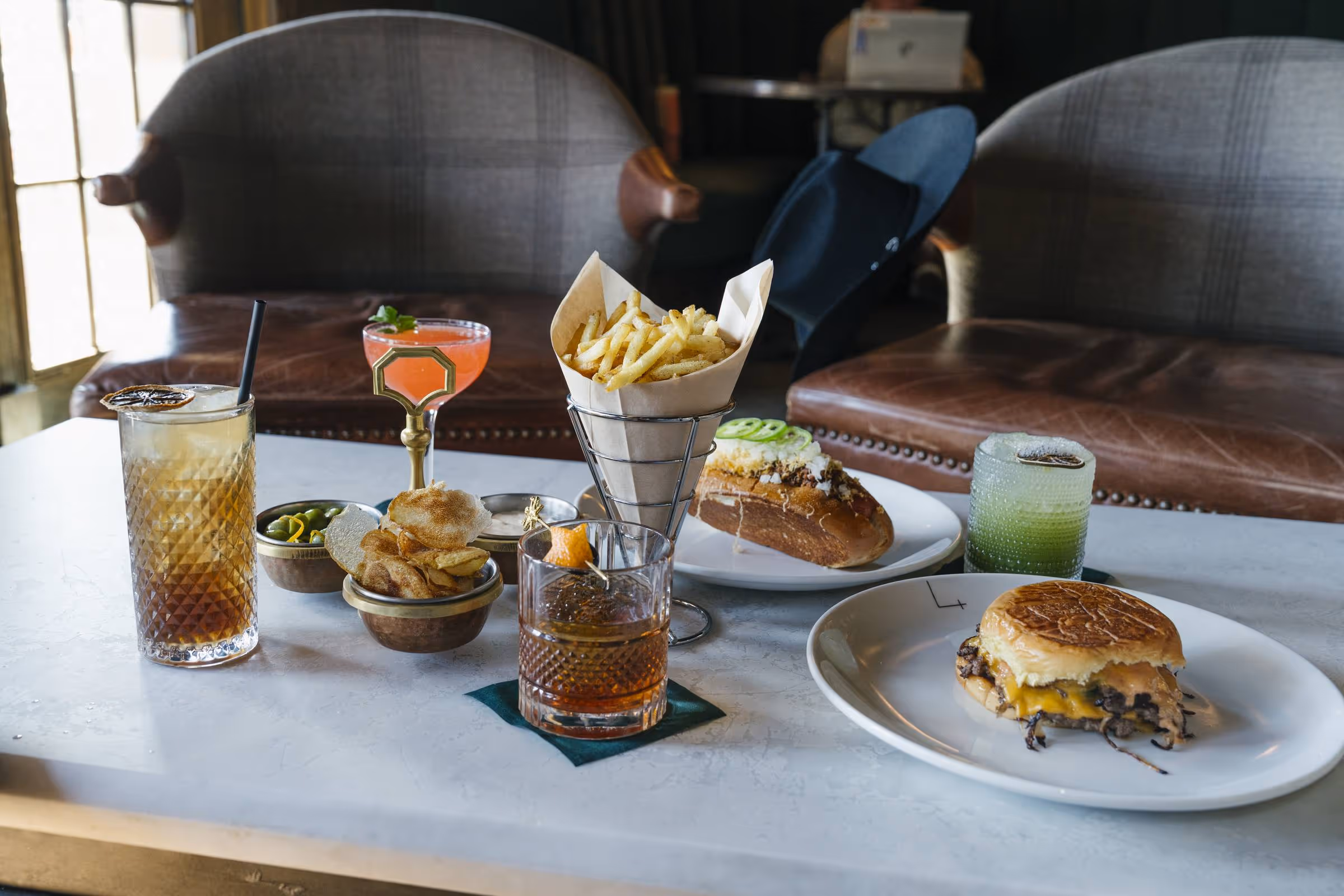 Bar spread with smash burger, chili dog, fries, and three cocktails on a marble table beside a leather club chair at Stewart's
