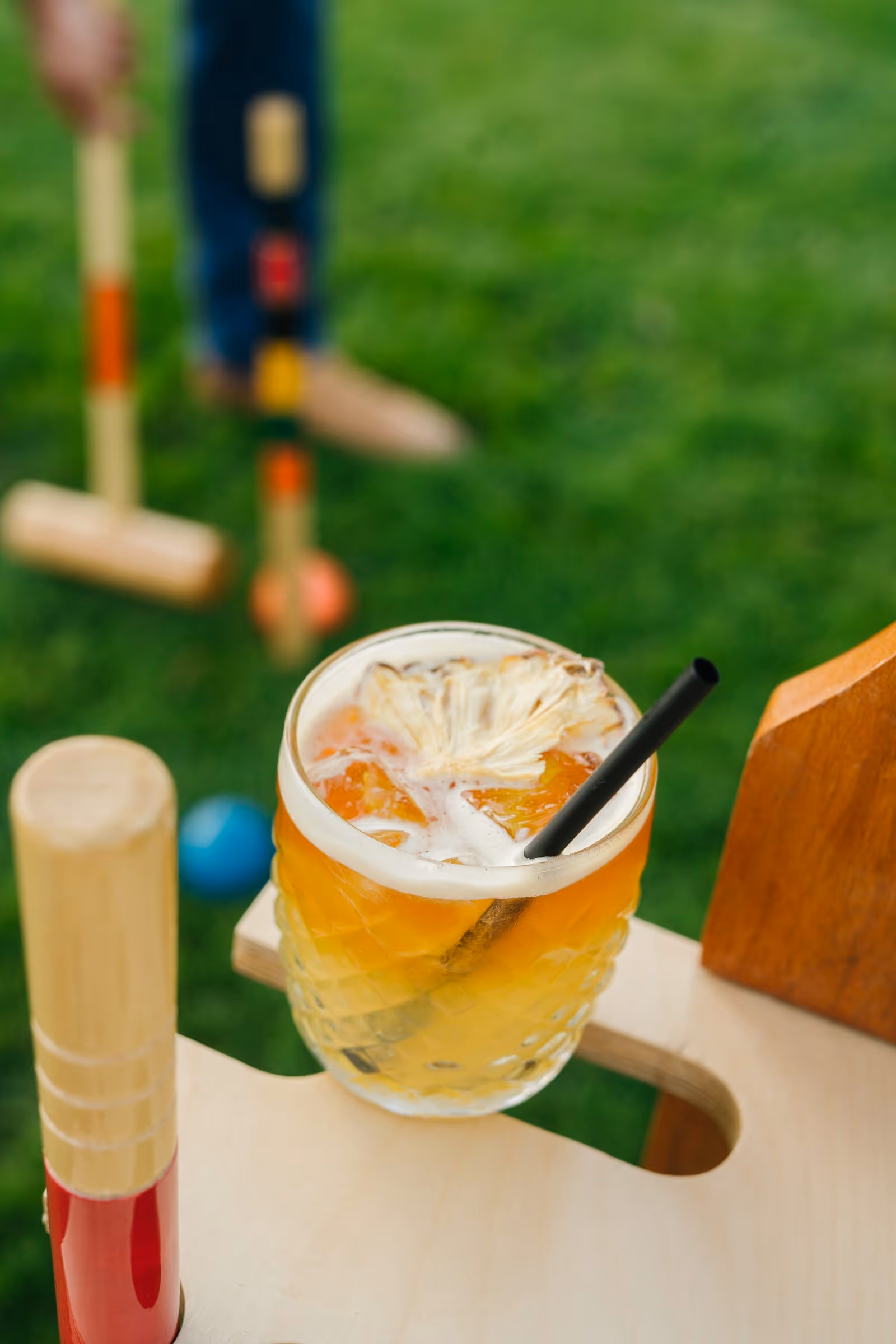 Tropical cocktail with dehydrated pineapple garnish on a wooden holder beside a croquet course at Stewart's