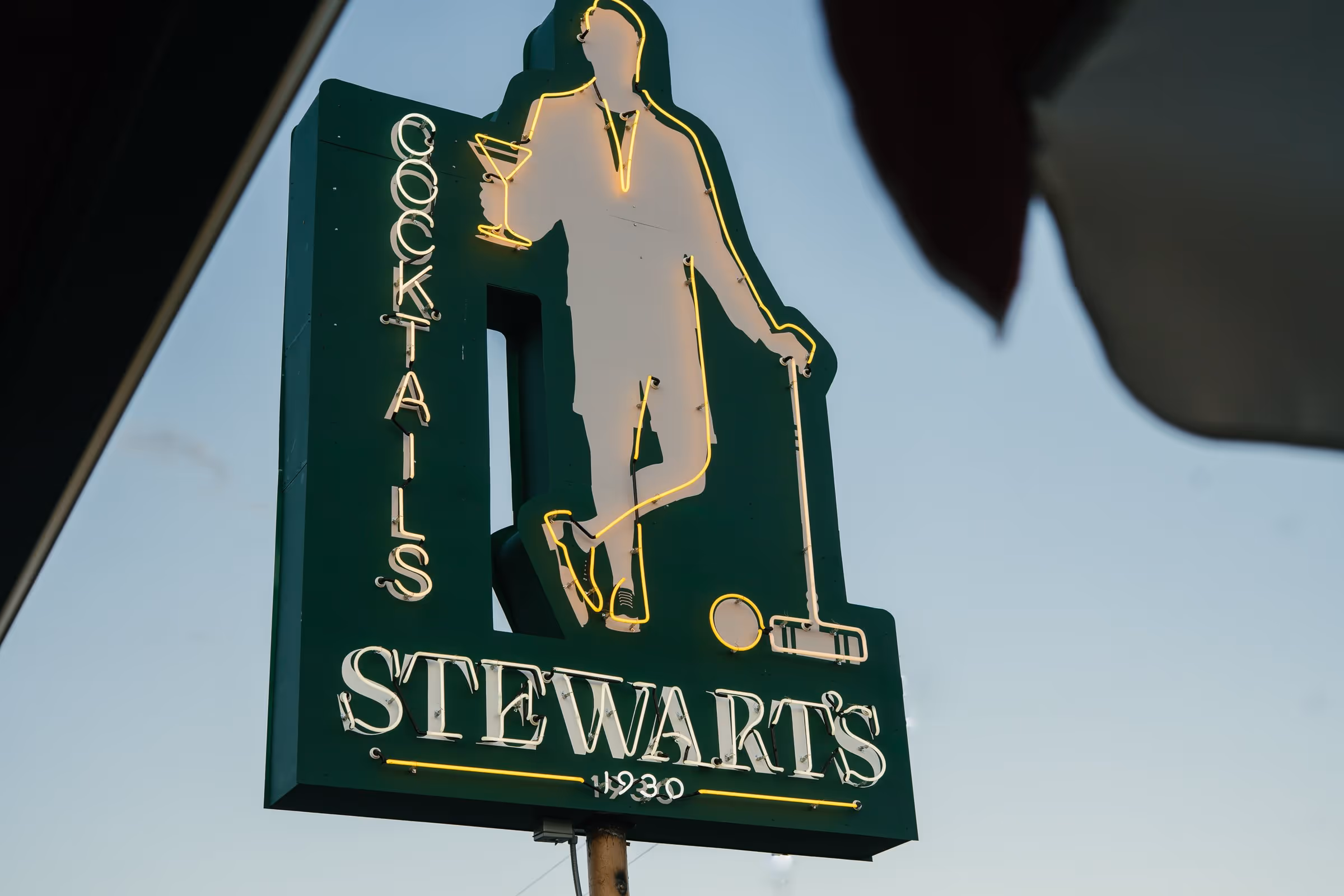 Illuminated vintage neon sign of a figure in a tuxedo holding a martini and croquet mallet above the Stewart's 1930 wordmark