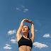 Young woman in black sports bra stretching arms above her head against a blue sky with scattered clouds.