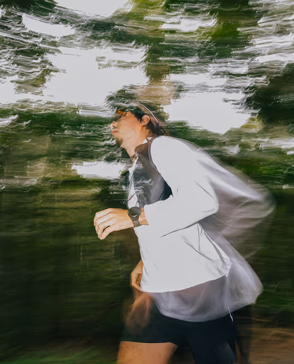 Man running outdoors with motion blur effect, wearing a white long-sleeve shirt, black shorts, sunglasses, and a visor.