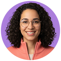 Portrait of a smiling woman with shoulder-length curly hair, wearing glasses and a salmon-pink zip-up sweater.