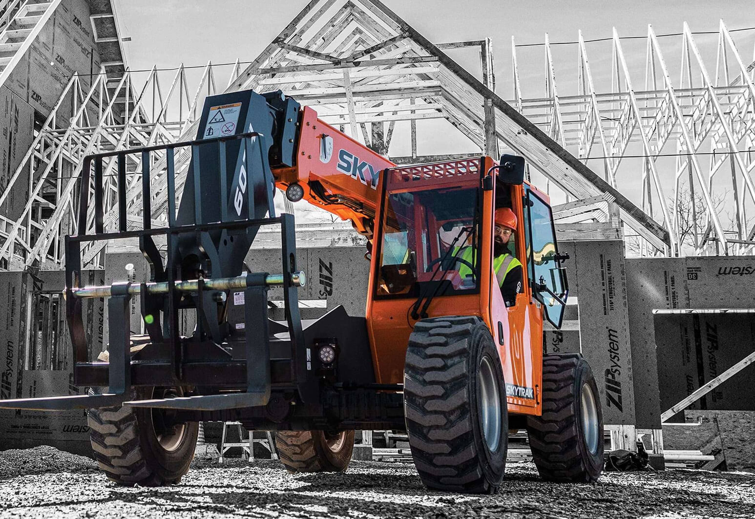 Orange SkyTrak forklift operated by a worker wearing a safety vest and helmet at a wooden house construction site.