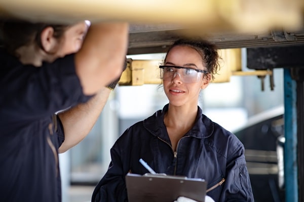 A mechanic stands under a car on a lift while holding an inspection checklist