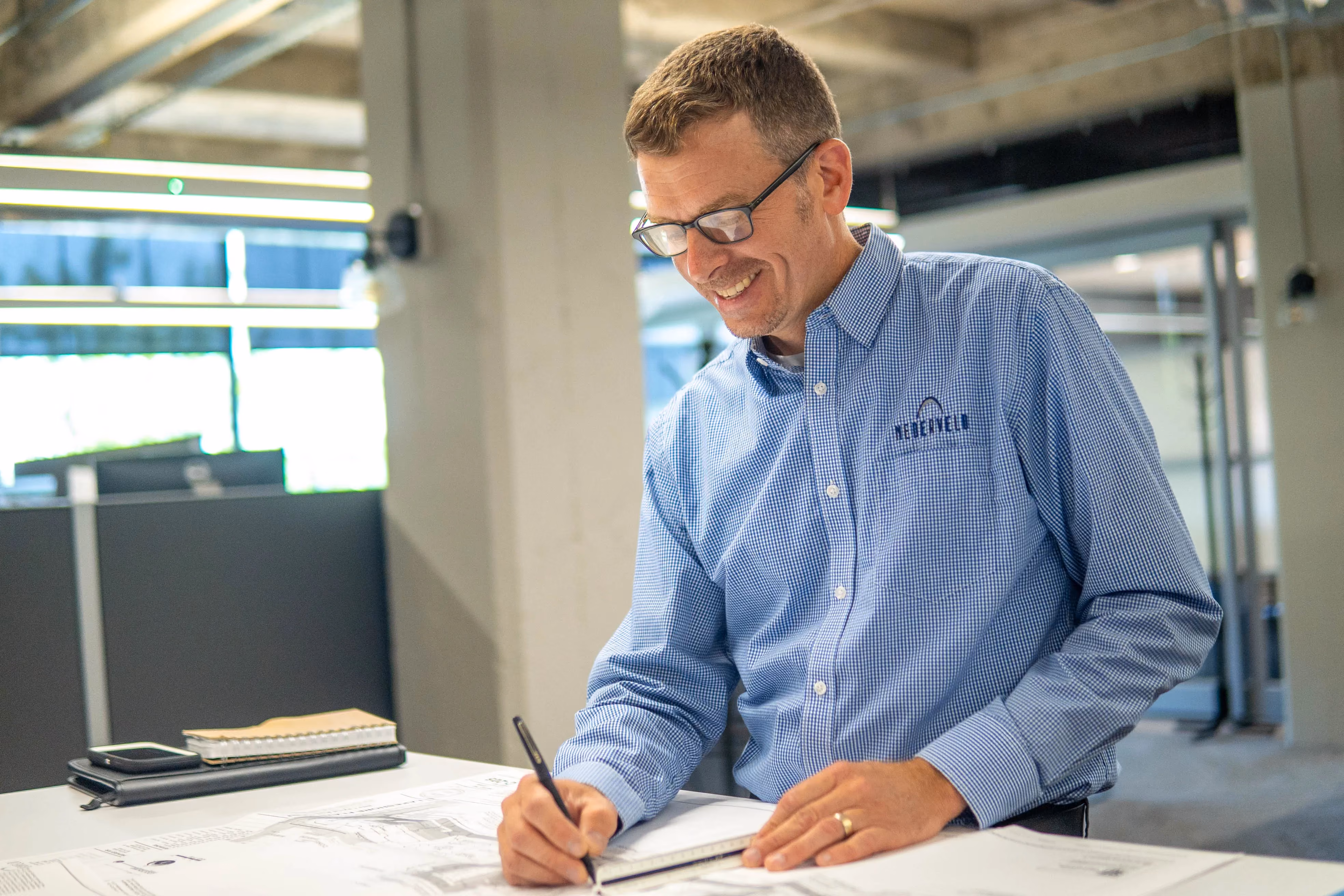 Man wearing glasses and a blue checkered shirt with a company logo smiling and working on architectural blueprints at a desk in an office.