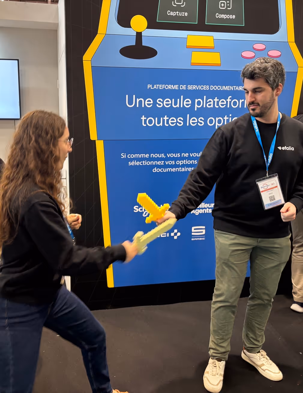 Deux personnes se tiennent dans un stand avec un jeu vidéo rétro en arrière-plan, chacune tenant une épée en mousse