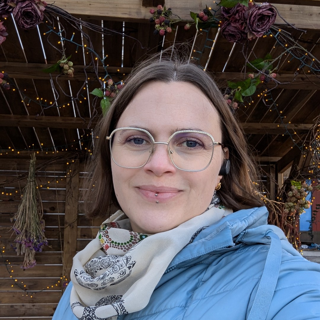 Photo of Emma from the shoulders up. She's outside and wear a blue jacket, light scarf, and glasses. She's looking into the camera and smiling. 