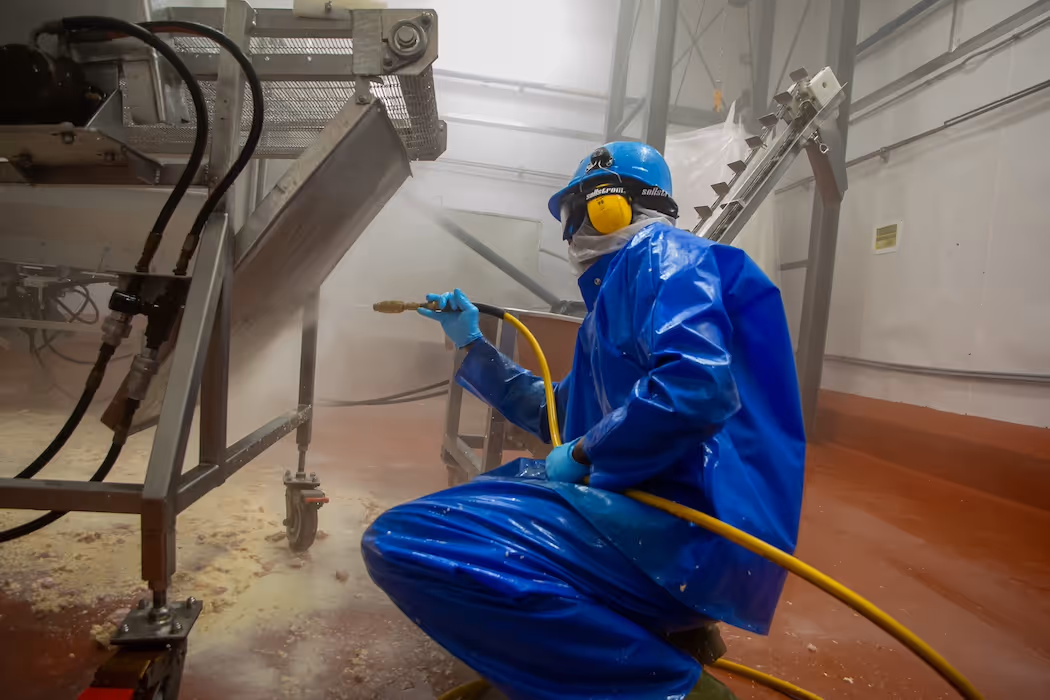 Worker in blue protective gear spraying and cleaning industrial machinery inside a factory.