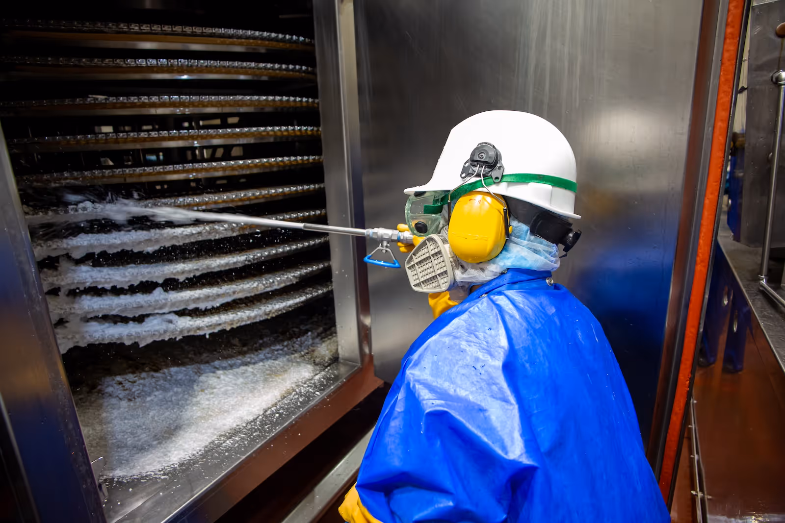 Worker in protective gear using a high-pressure spray to clean industrial equipment with metal racks.