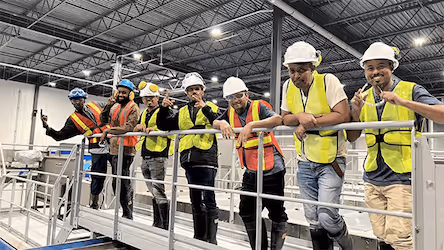 Seven construction workers wearing safety helmets and high-visibility vests standing on an indoor platform, smiling and posing for a photo.