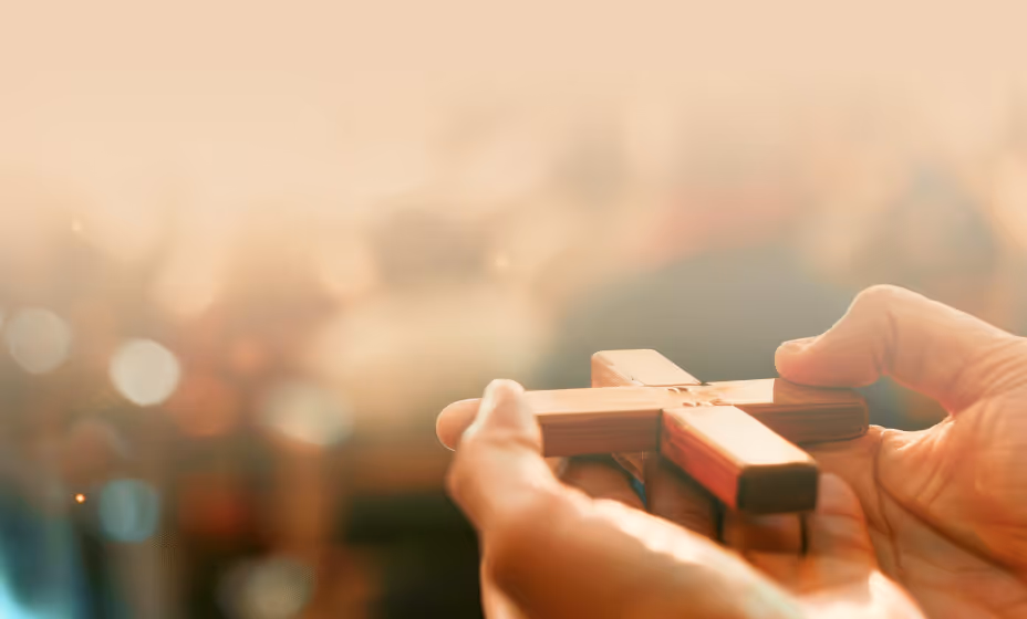 Hands holding a small wooden cross against a warm, blurred background, symbolizing personal prayer and spiritual devotion.