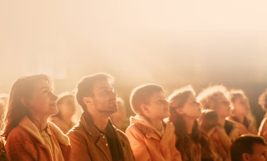 People stadning together looking upward in a warmly lit worship service, conveying attentive listening, community, and shared faith.