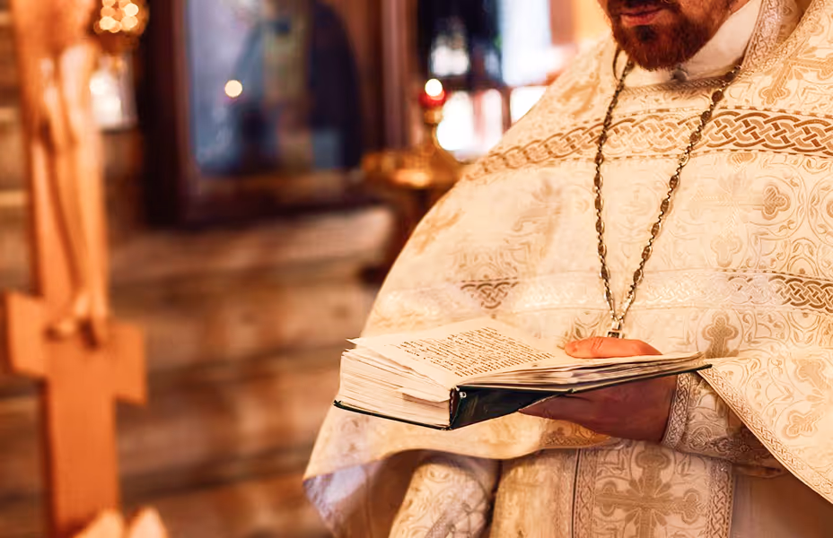 Orthodox priest in ornate vestments holding an open liturgical book during the Divine Liturgy, illustrating the proclamation of Scripture and the sacramental life of the Church.