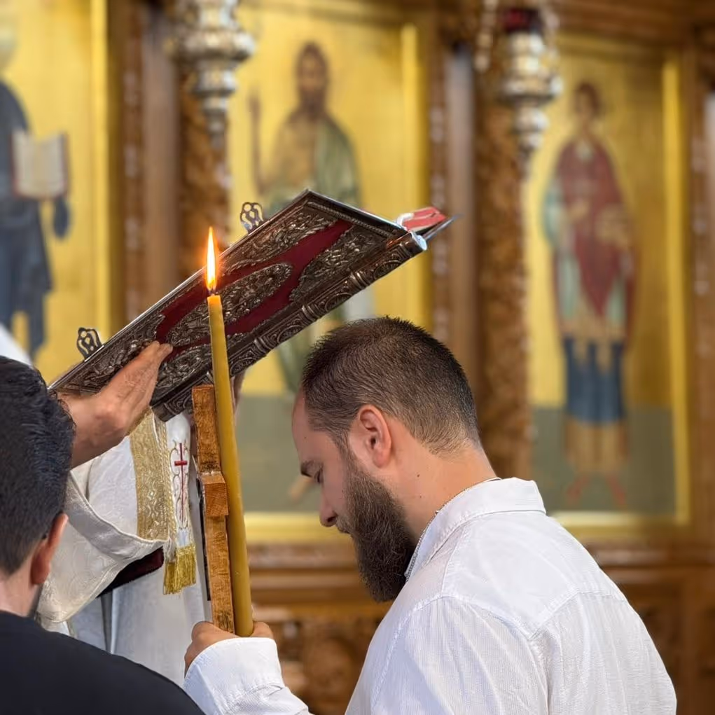Man with a beard holding a lit candle and bowing his head during a religious ceremony in a church with icon paintings in the background.