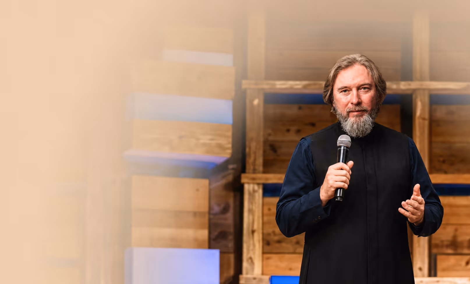 Father Viktor in black holding a microphone, speaking in front of a wooden pallet backdrop.