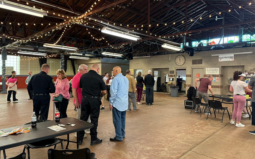 People gather and talk inside a large hall with string lights, tables, and chairs; signs for restrooms and farmers market are visible on the wall.