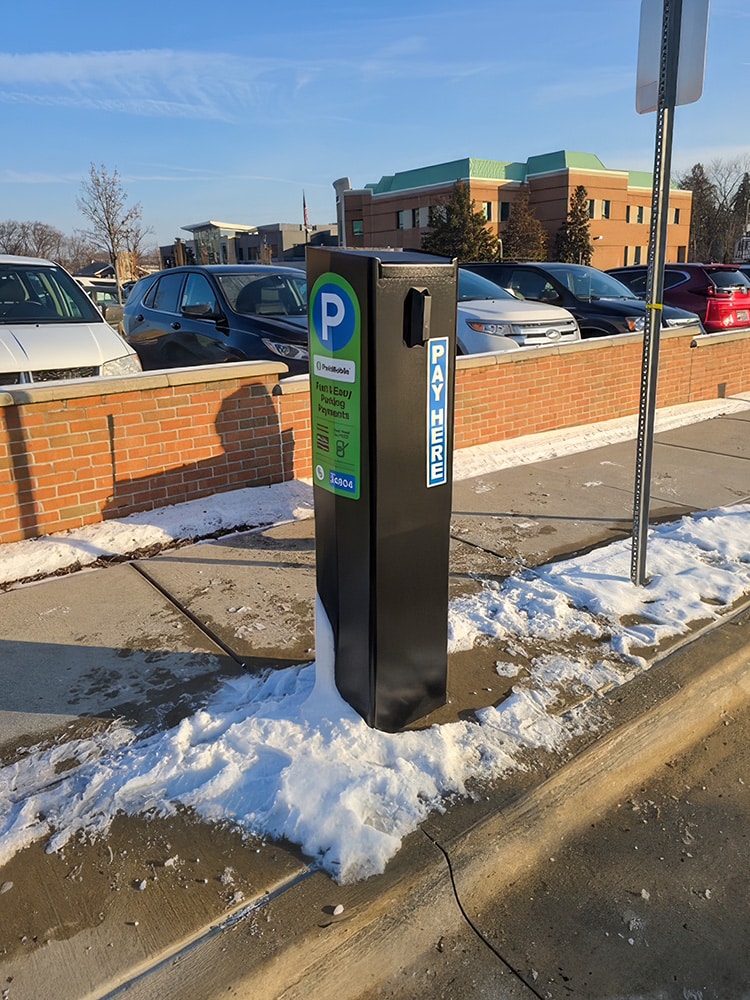A black parking payment kiosk stands on a snowy sidewalk next to parked cars, with a "Pay Here" sign and instructions visible.