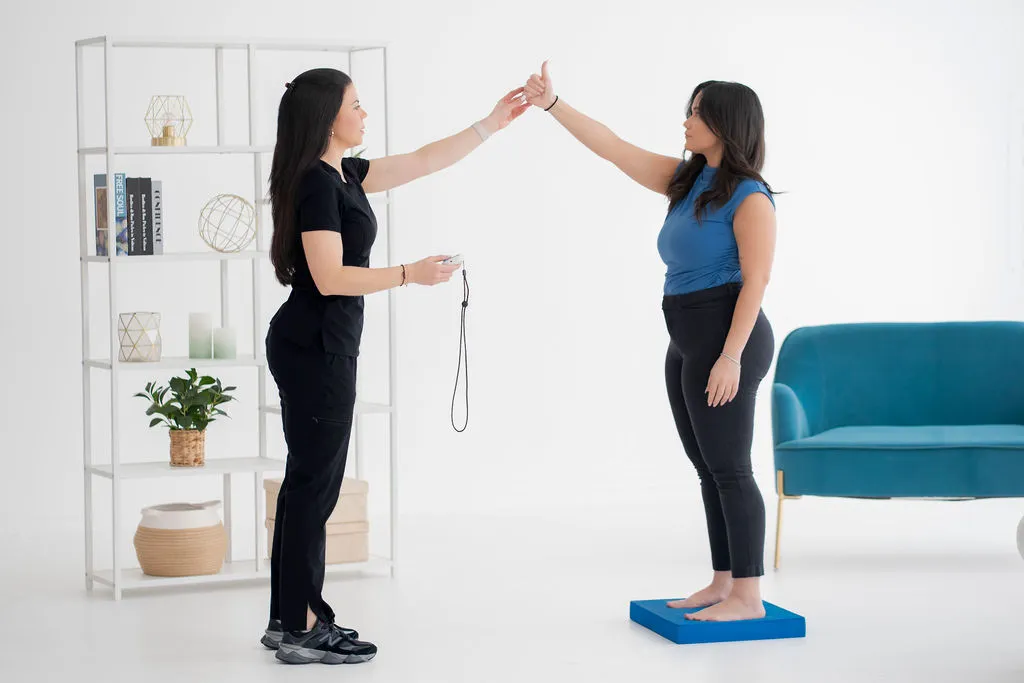 Medical professional conducting a balance assessment with a patient standing barefoot on a blue balance mat, both with arms raised, in a bright clinic setting with modern furniture.