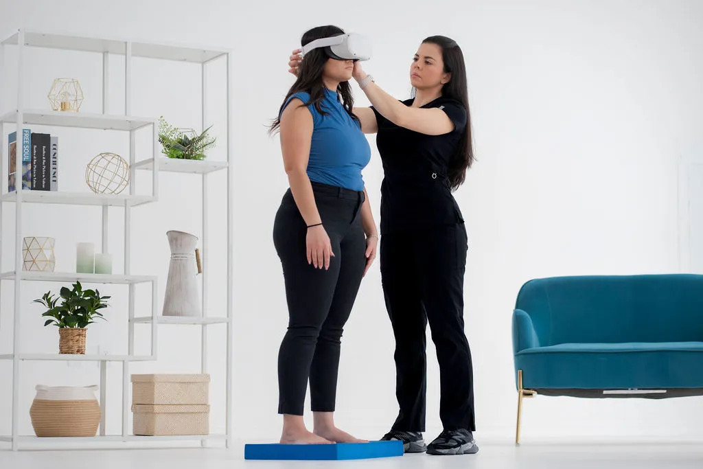 Medical professional adjusting a VR headset on a patient standing on a balance platform during a vestibular assessment or therapy session in a bright clinic setting, indicating the use of virtual reality for balance disorders.