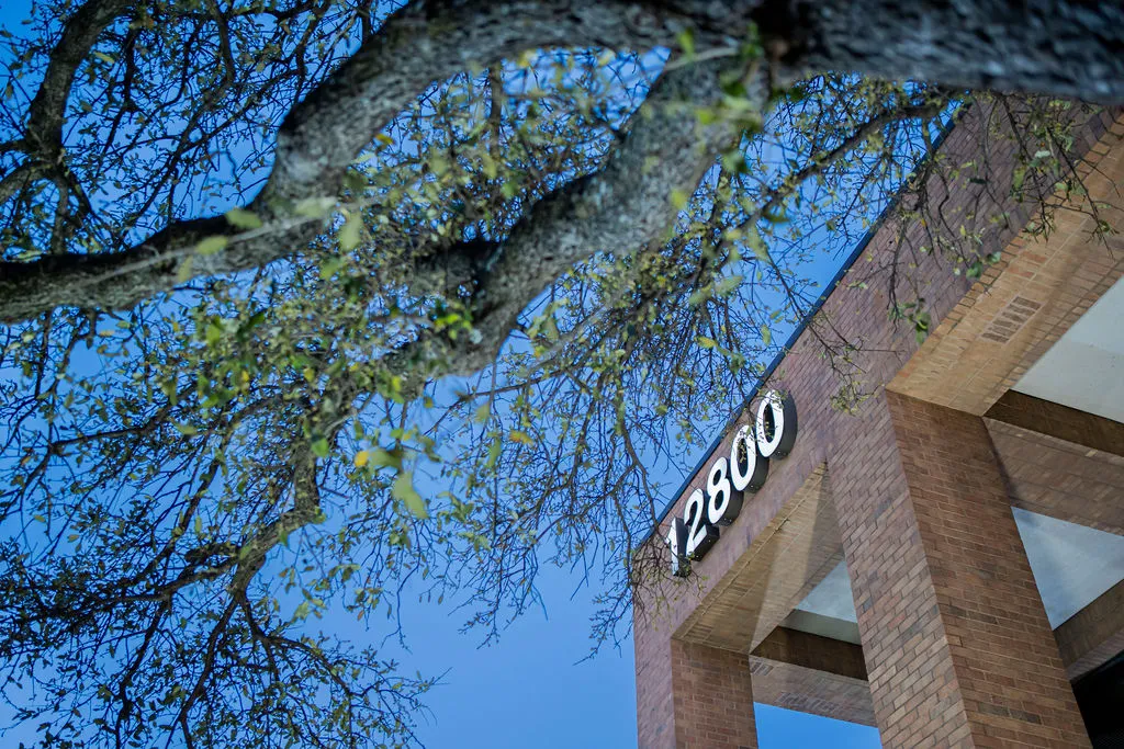 Exterior view of a modern brick building featuring the number '12800' on its facade, partially visible through tree branches against a blue sky, indicating the clinic's location.