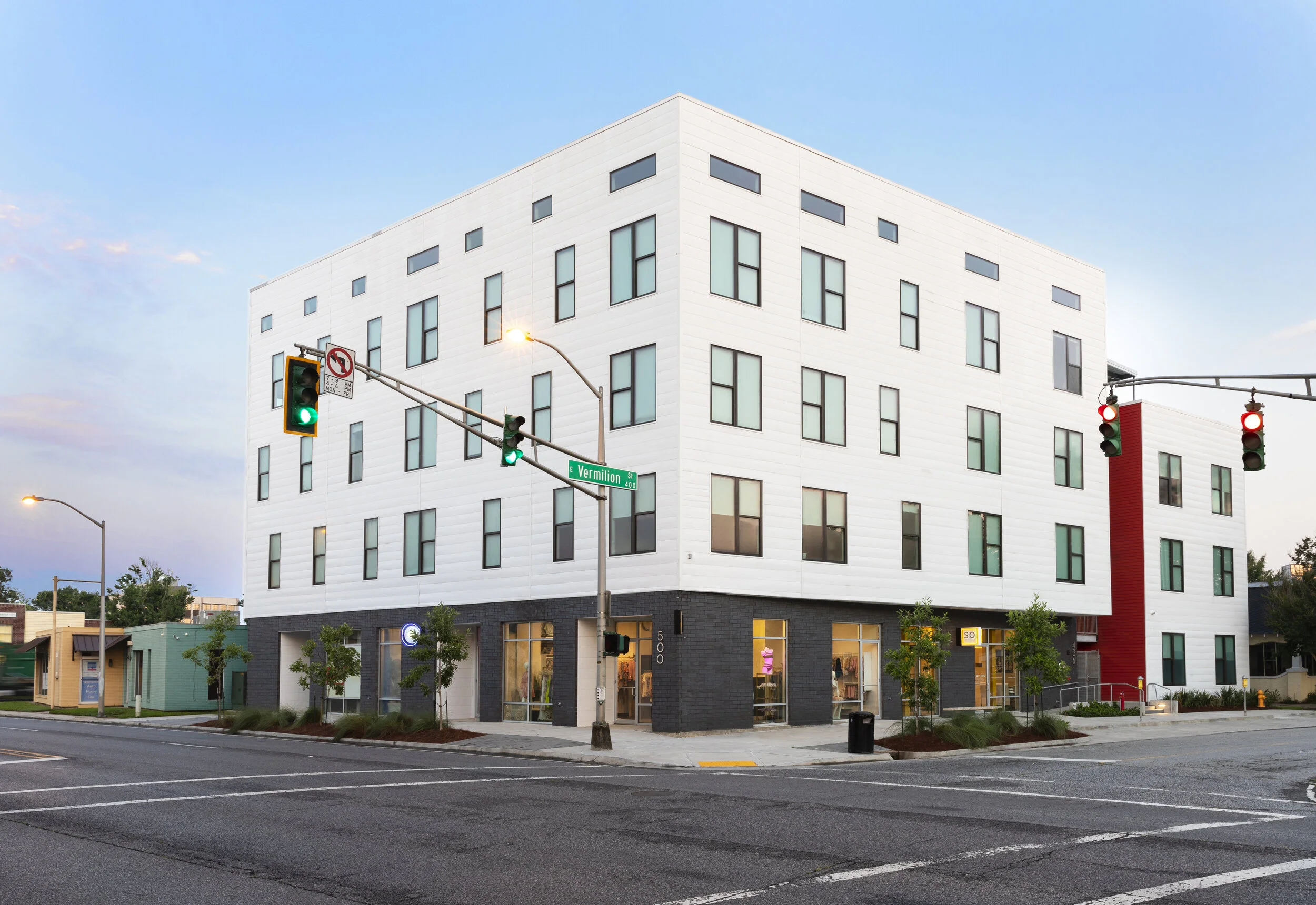 Modern four-story residential building on a corner with street signs and traffic lights at a clear dusk.
