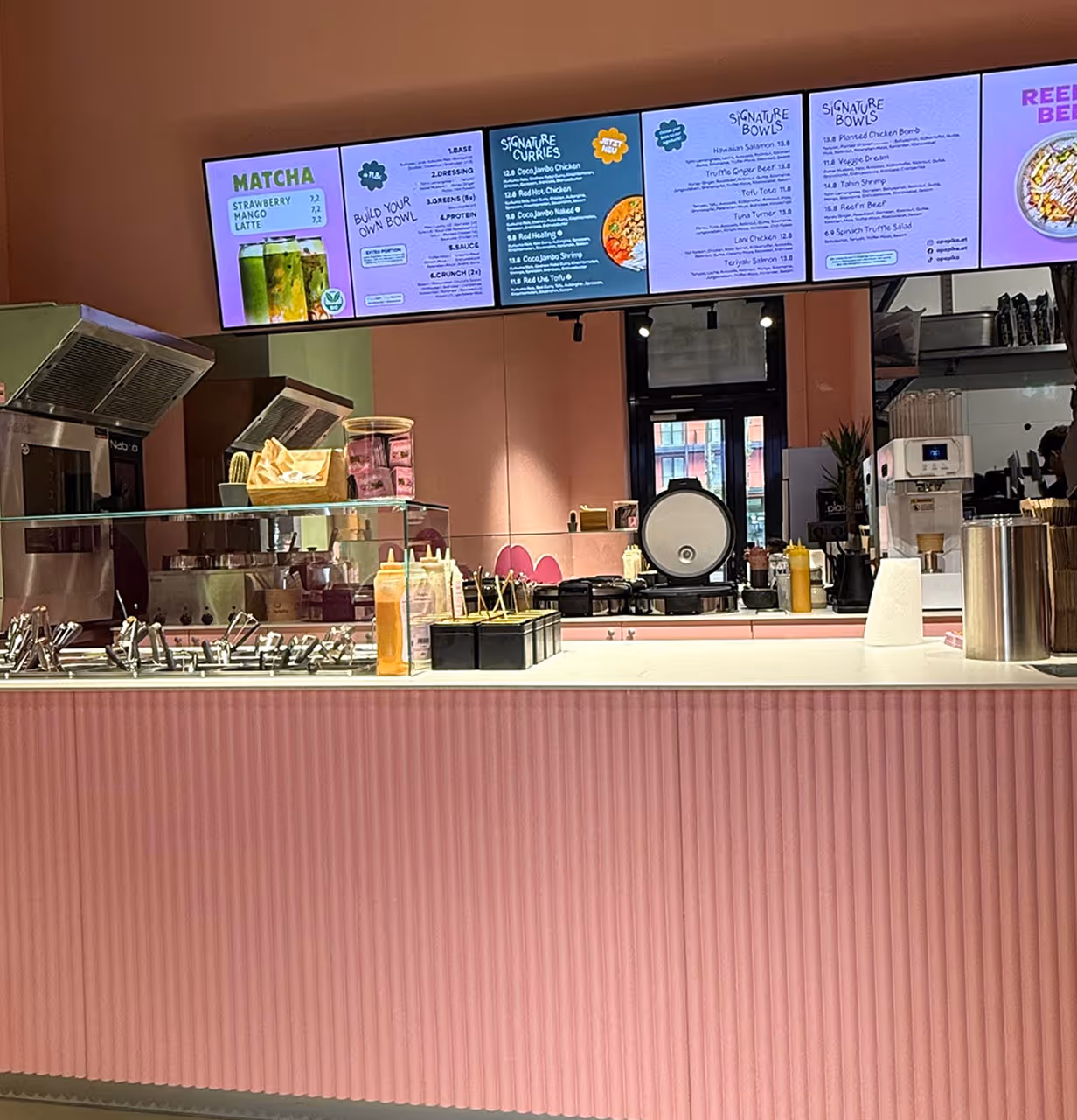 Eine moderne Café-Theke in Pink zeigt eine digitale Speisekarte mit Speisen und Getränken. Speisen und Utensilien sind übersichtlich angeordnet und strahlen eine einladende Atmosphäre aus.