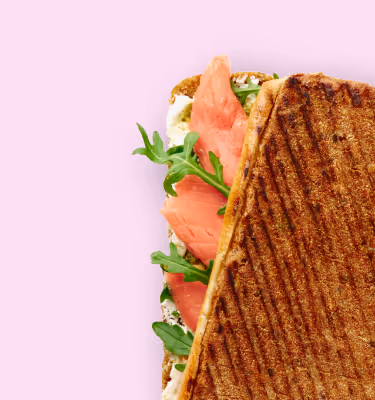 Gegrilltes Sandwich mit Räucherlachs und Rucola auf Frischkäsebrot vor rosa Hintergrund.