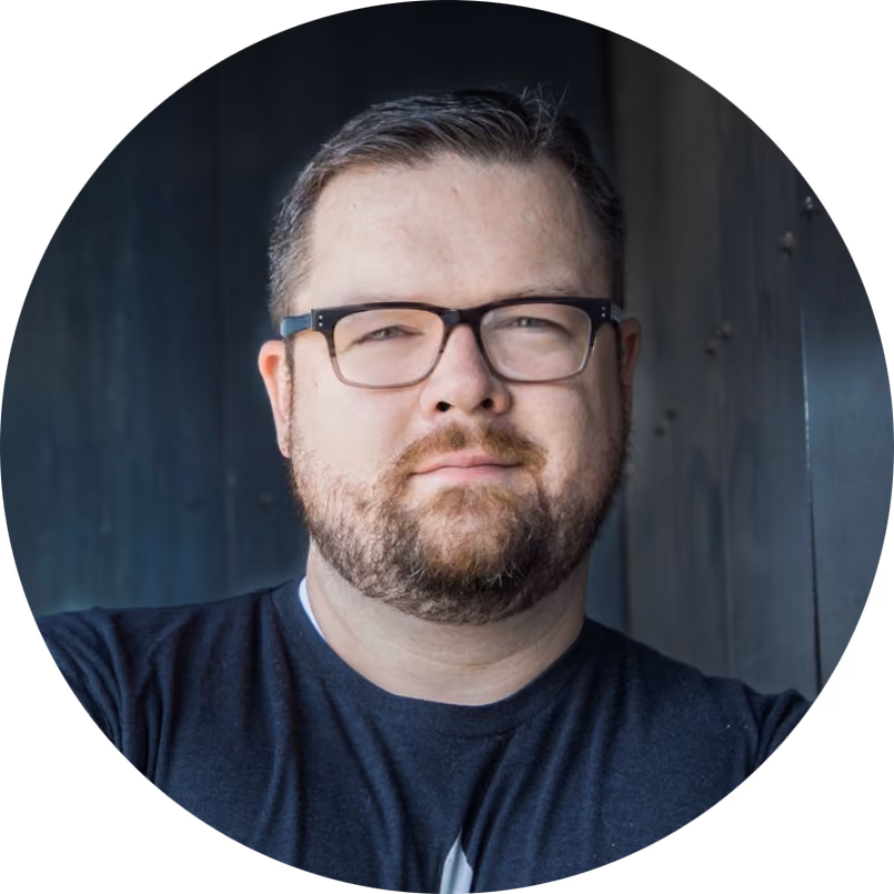 Bearded man wearing glasses and a dark shirt looking at the camera with a neutral expression.