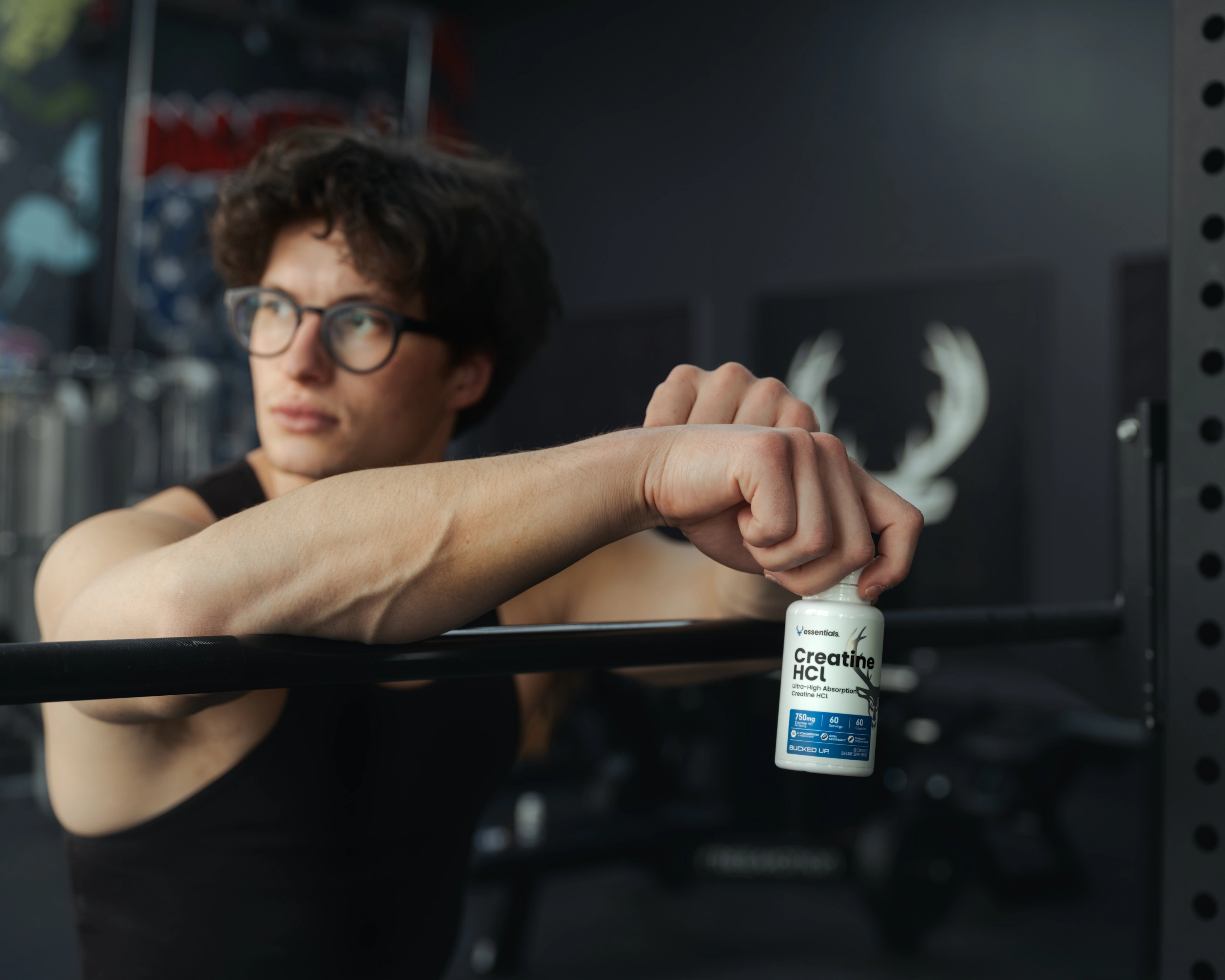 Man wearing a tank top holding Creatine HCl by the lid in a gym.