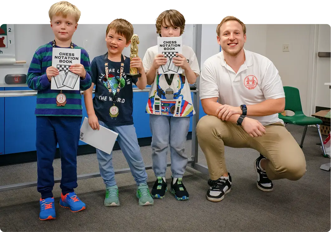 A chess instructor kneels beside three young students who are smiling and proudly displaying their chess notation books, medals, and a trophy at Silicon Valley Chess.